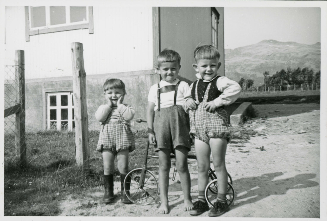 Tre barn med korte bukser smiler sjenert til fotografen. 
