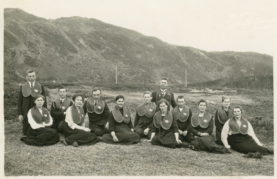 Losjen i Saursfjord, en gruppe unge menn og kvinner sitter pent antrukket utendørs, med losjekraver på. 